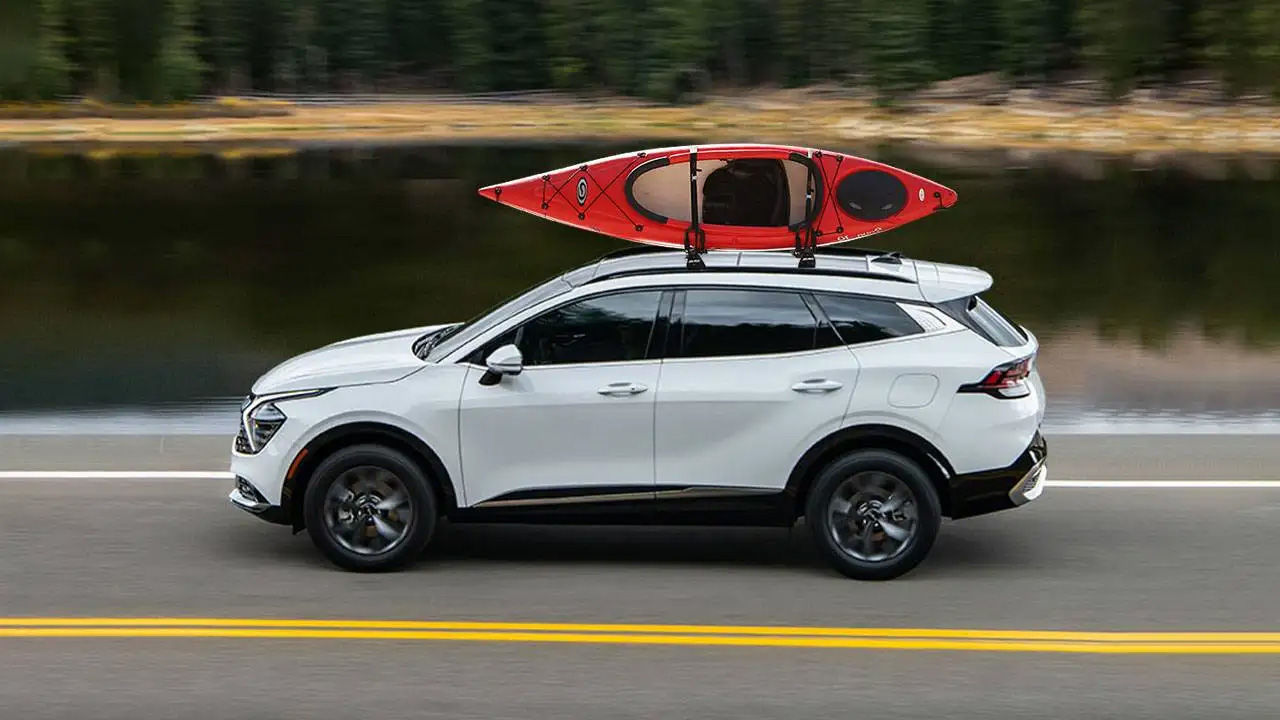 A white Kia Sportage SUV with a red kayak mounted on the roof, driving along a road next to a body of water.
