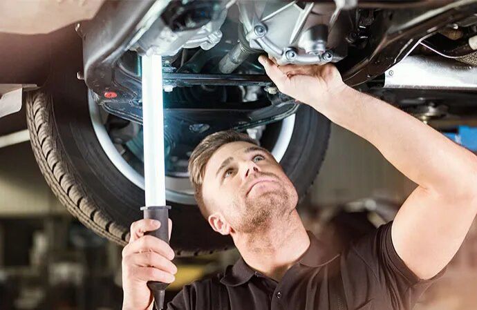 A mechanic underneath a car lift using a work light to inspect the vehicle's undercarriage.
