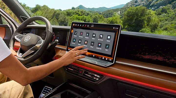 A hand interacts with the large central infotainment screen, which displays the home menu with various apps and controls.