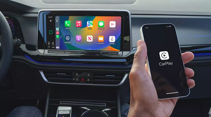 Close-up of the infotainment screen displaying the Apple CarPlay interface, with a hand holding a phone showing the CarPlay connection screen.