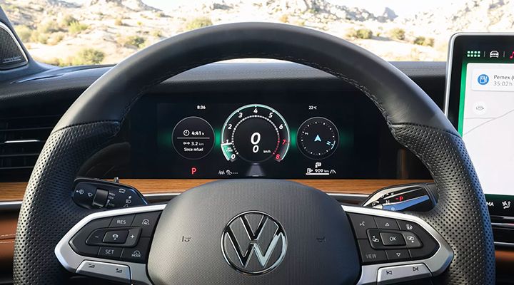 View through the steering wheel, showing the driver's digital instrument cluster with the tachometer, speedometer, and driving information. The steering wheel is leather-wrapped and the interior features wood trim.