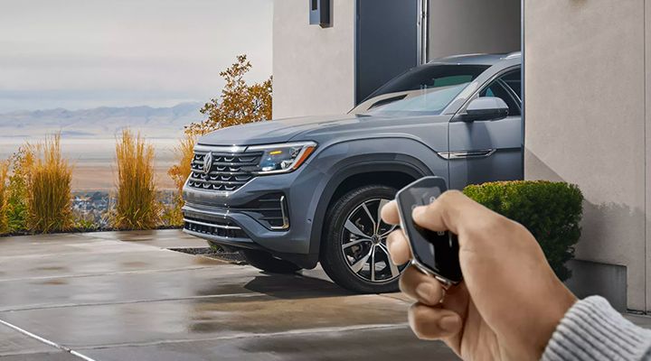 Hand using a key fob to unlock or remotely start a grey Volkswagen Atlas or Atlas Cross Sport parked in front of a house.