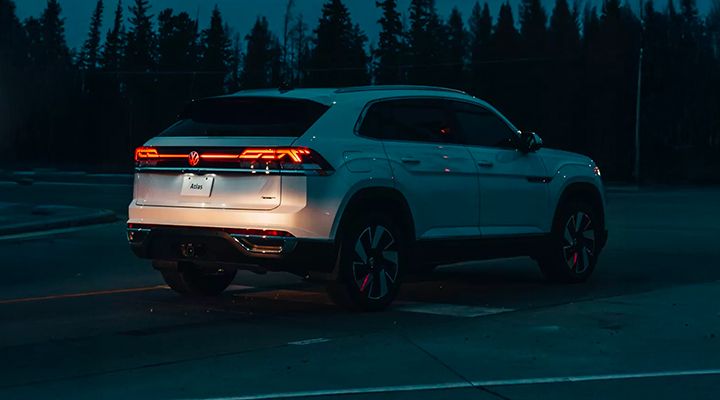 Night rear view of a white Volkswagen Atlas or Atlas Cross Sport in a parking lot, highlighting the LED taillight bar and illuminated Atlas logo.