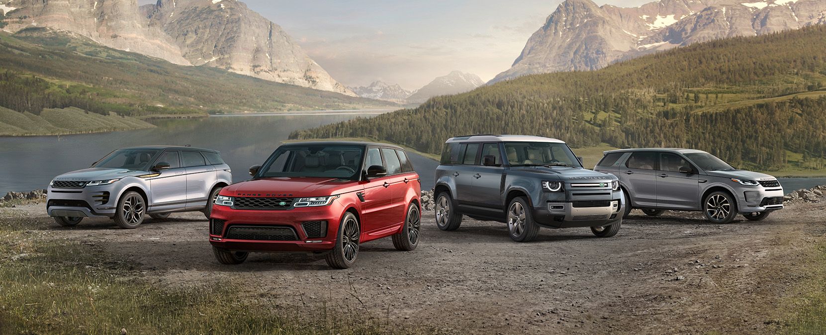 Four Land Rover SUVs – a silver Range Rover Evoque, a red Range Rover Sport, a blue Land Rover Defender, and a grey Range Rover Velar – parked on a gravel surface with a lake and mountains in the background.