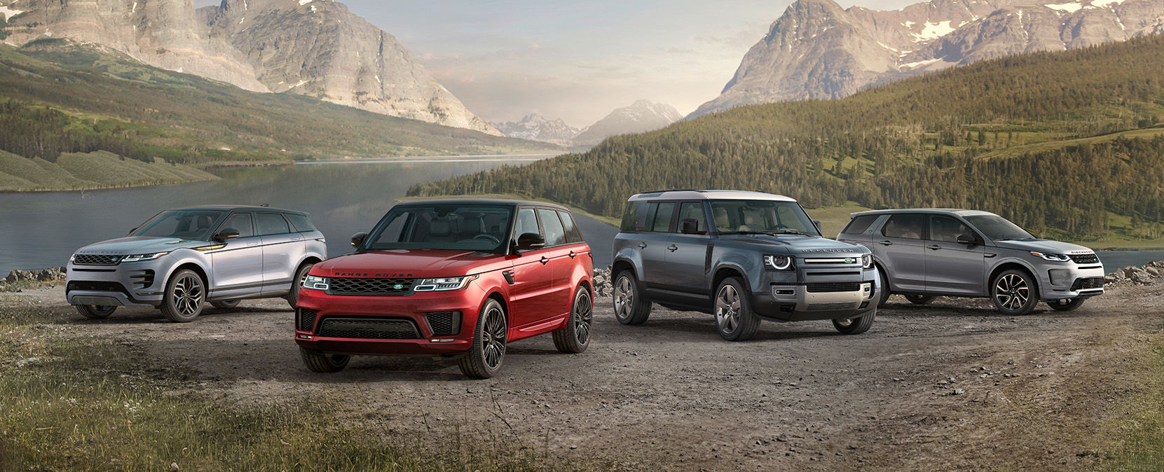 Four Land Rover SUVs – a silver Range Rover Evoque, a red Range Rover Sport, a blue Land Rover Defender, and a grey Range Rover Velar – parked on a gravel surface with a lake and mountains in the background.