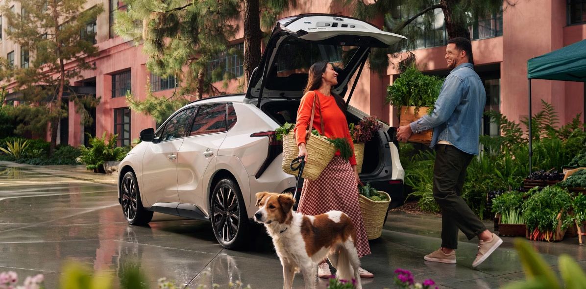 A couple loading baskets and plants into the open trunk of a white Toyota bZ4X, with a dog on a leash in the foreground.