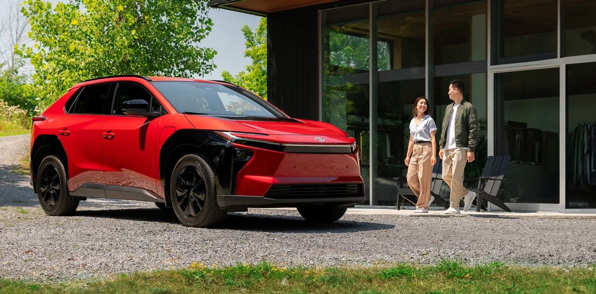 A red Toyota bZ4X parked in front of a modern glass house, with a couple walking nearby.