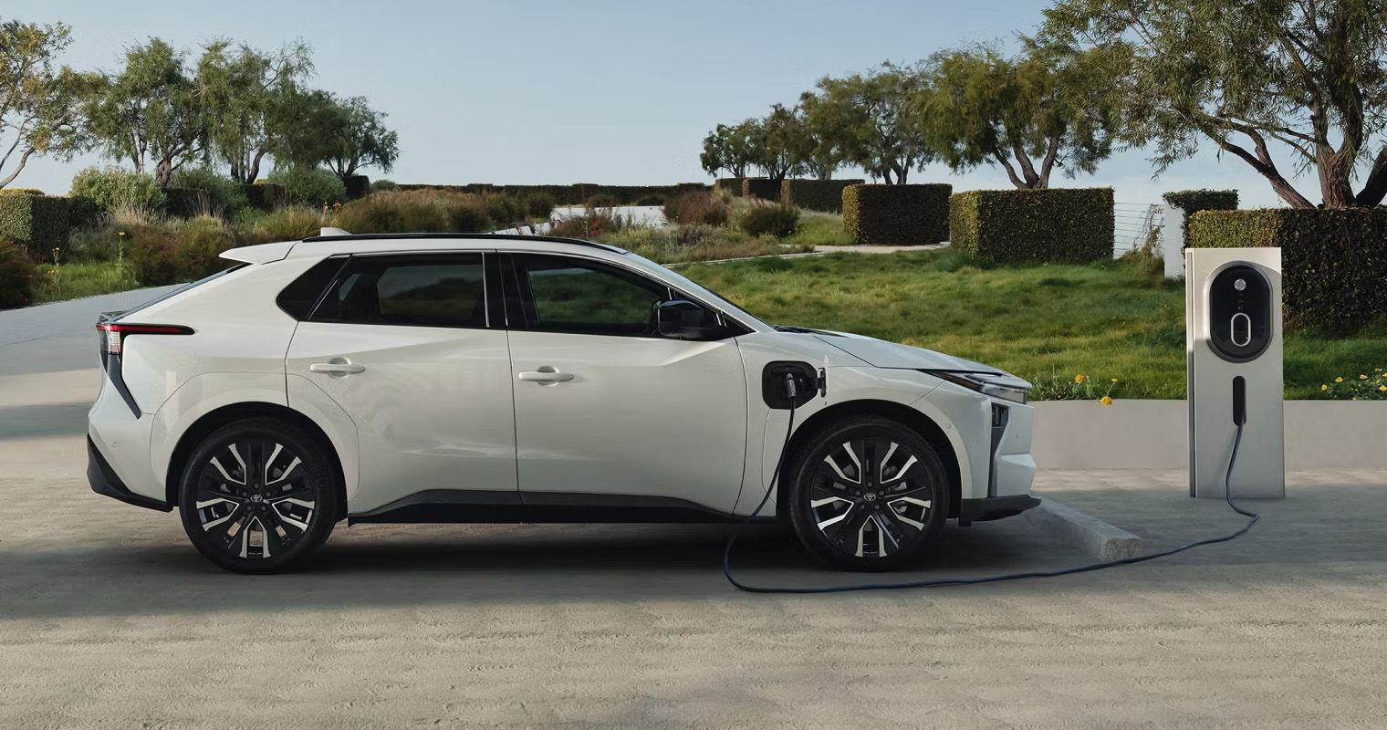 Side profile of a white Toyota bZ4X plugged into an electric charging station in a park setting.