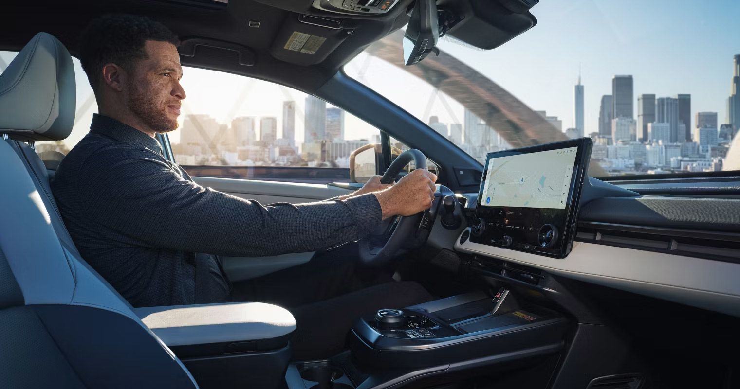 Interior view of a driver behind the wheel of a Toyota bZ4X, showing the large central touchscreen and a city view.