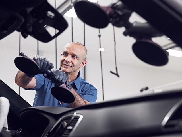 A male technician, who appears to be of Middle Eastern or South Asian descent, carefully installs a new windshield on a vehicle using professional suction cup tools.