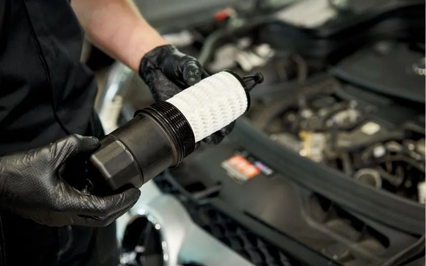 A mechanic wearing black gloves holds a new white car oil filter over an engine bay.