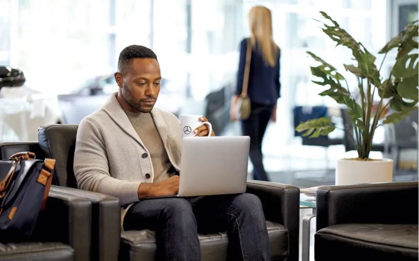 A man sitting in a dealership lounge uses his laptop while holding a mug.
