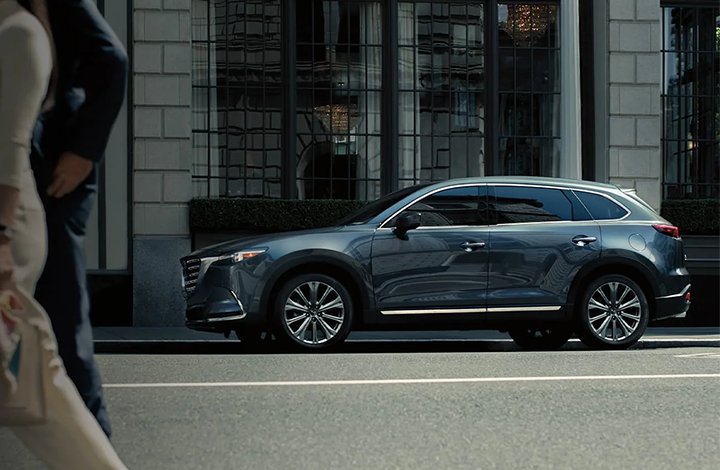 A grey Mazda SUV parked on a city street in front of a building with large windows.