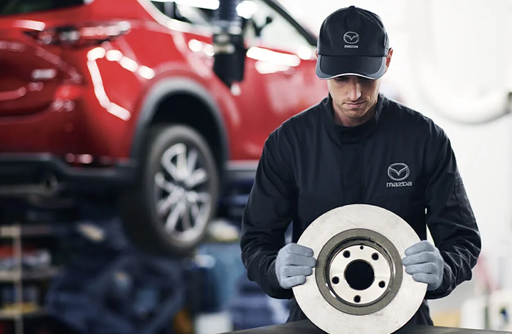 A Mazda technician in a dark uniform and cap holding a silver brake rotor.
