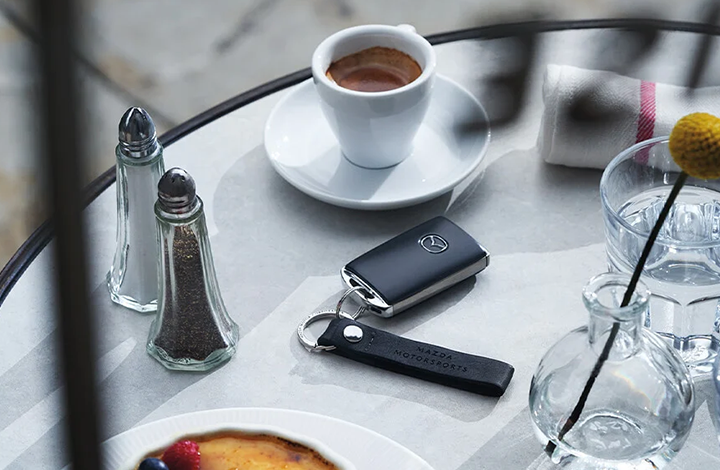 A Mazda key fob with a leather "Mazda Motorsports" keychain on a cafe table next to a cup of espresso.