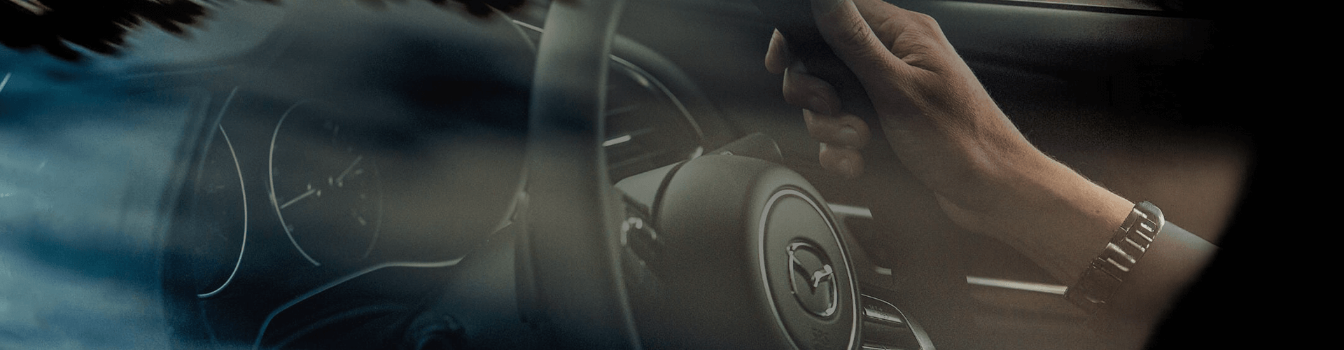 A close-up, cinematic shot of a driver’s hand on a Mazda steering wheel, showing the dashboard and logo through a glass reflection.