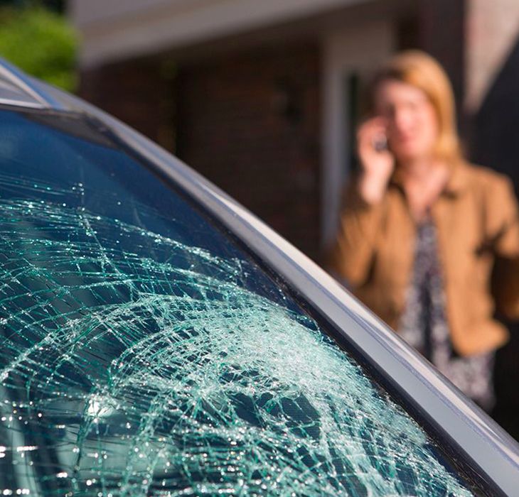 A close-up shot of a severely shattered car windshield with a spiderweb crack pattern. In the blurred background, a woman stands outside a house talking on a cell phone.