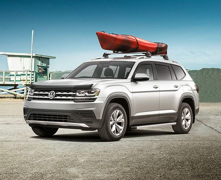 A silver Volkswagen Atlas parked on a gravel surface near a beach. It features a black hood deflector and a red kayak mounted on its roof rack.