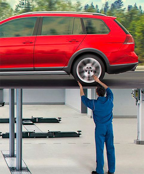 A red Volkswagen Golf Alltrack on a hydraulic service lift in a bright, modern garage. A technician in blue coveralls is positioned underneath, reaching up toward the rear wheel.