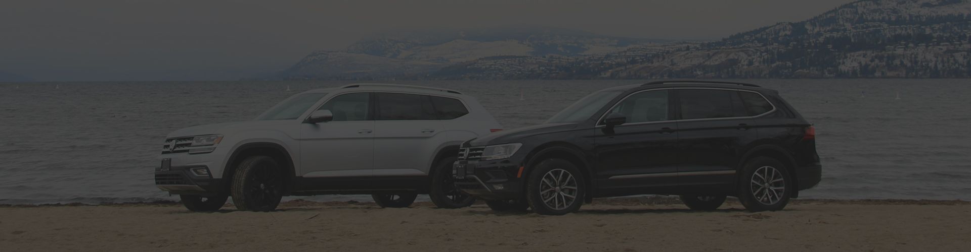 A wide-angle, low-light photograph of a white SUV and a black SUV parked side-by-side on a sandy beach. A large body of water and snow-capped mountains are visible in the background under an overcast sky.