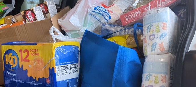 A close-up of a car trunk packed with groceries and household items, including a 12-pack of Kraft Dinner, a blue reusable bag, a large stack of diapers, and a package of Tim Hortons soup.