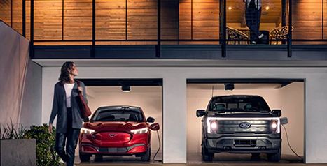 A red Mustang Mach-E and a silver F-150 Lightning charging overnight in a modern two-car garage under a lighted balcony.