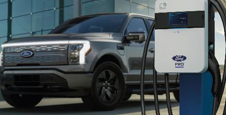 A close-up of a Ford Pro commercial charging station with a silver F-150 Lightning blurred in the background.