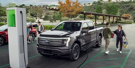 A silver Ford F-150 Lightning parked at a public charging plaza while people interact nearby.