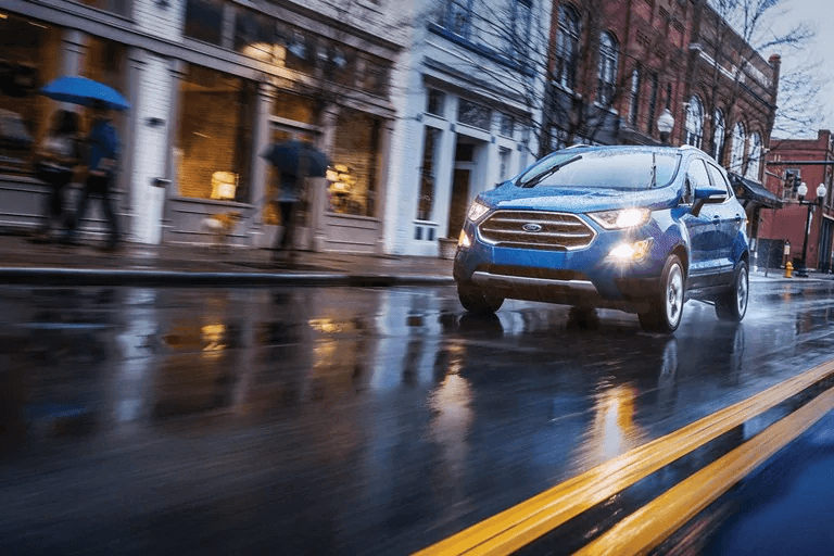 Un VUS bleu Ford EcoSport circulant en ville sur une chaussée asphaltée et mouillée par la pluie, reflétant les lumières environnantes.