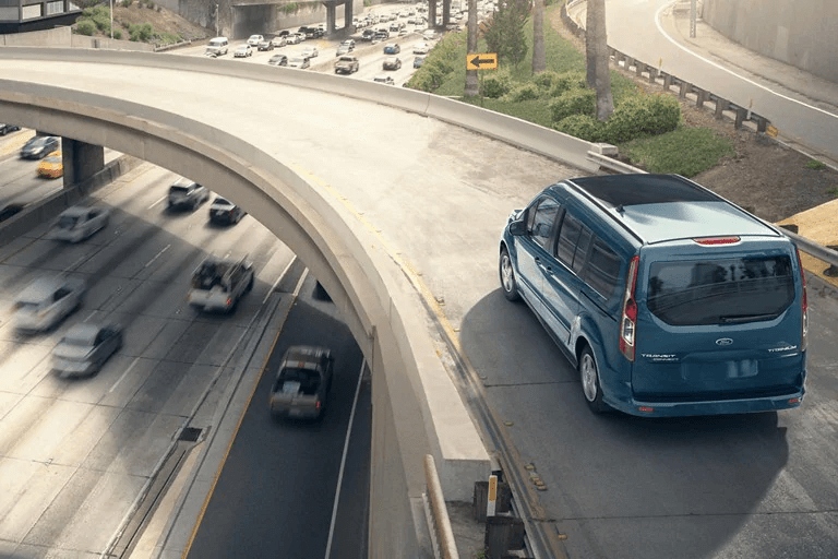 Vue en plongée d'une fourgonnette bleu sarcelle Ford Transit Connect circulant sur une rampe d'accès surélevée, surplombant une autoroute achalandée.
