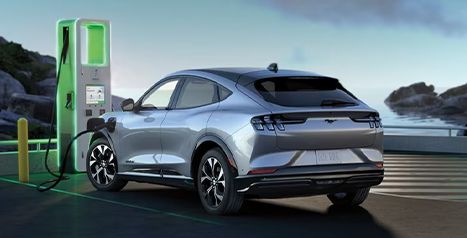 A silver Mustang Mach-E parked at a public charging station near the ocean, plugged into a green illuminated charger.