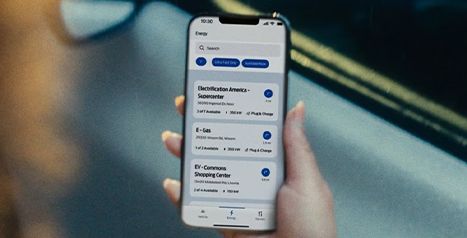 A hand holding a smartphone displaying a Ford app showing nearby electric vehicle charging station locations.