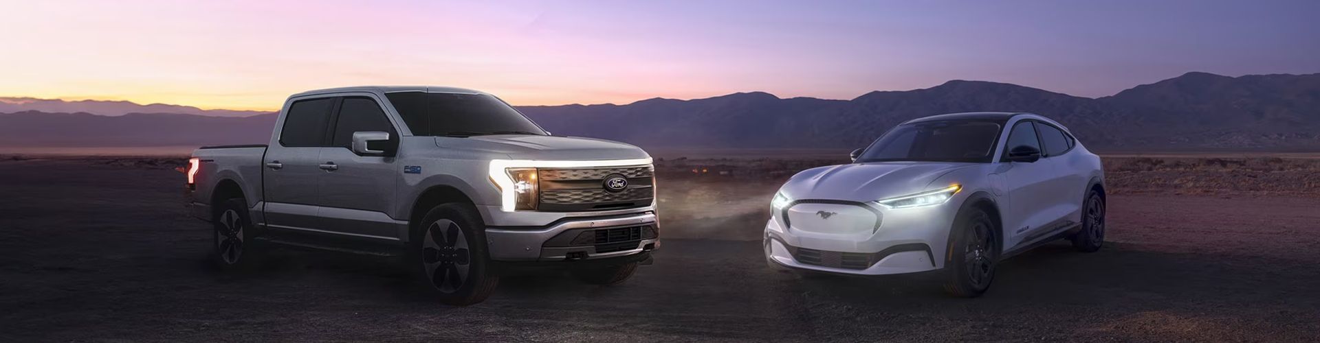 A Ford F-150 Lightning truck and a white Mustang Mach-E parked side-by-side in a desert landscape at twilight.