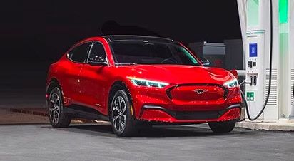 A red Ford Mustang Mach-E electric SUV plugged into a public fast-charging station at night.