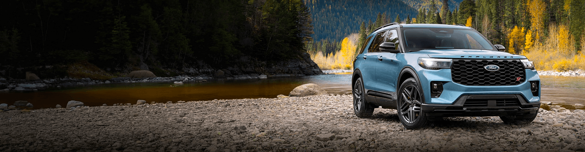 Un Ford Explorer ST bleu clair stationné sur une rive rocheuse au bord d'une rivière, avec une montagne boisée en arrière-plan.