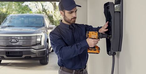 A professional technician using a power drill to install a Ford Charge Station Pro on a garage wall, with a silver F-150 Lightning parked in the background.
