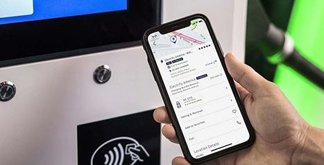 A hand holding a smartphone displaying the Electify America app interface in front of a charging station terminal with a contactless payment symbol.
