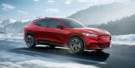 A red Mustang Mach-E driving on a snowy mountain road with bright blue skies in the background.