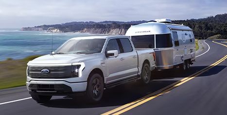 A silver Ford F-150 Lightning electric pickup truck towing a classic silver Airstream trailer along a scenic coastal road.