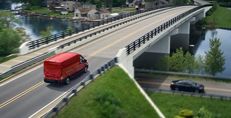 An aerial view of a red Ford E-Transit electric van driving across a bridge over a river in a suburban area.