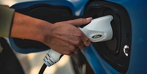 Close-up of a person's hand plugging a Ford electric vehicle charging cable into a blue car's charging port.