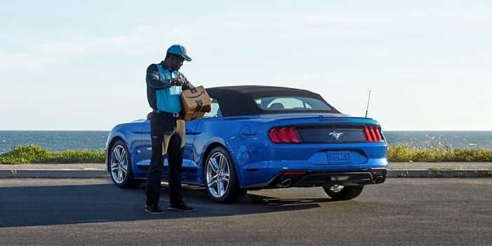 An Amazon delivery driver in uniform placing a package next to a blue Ford Mustang convertible parked by the ocean.