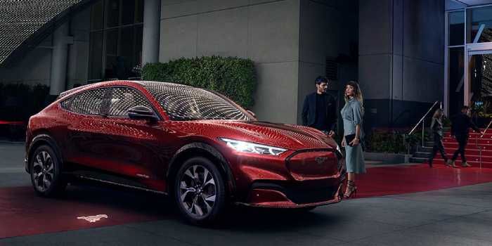 An elegant couple standing next to a glossy red Ford Mustang Mach-E electric SUV, parked in front of a modern building's illuminated entrance in the evening.