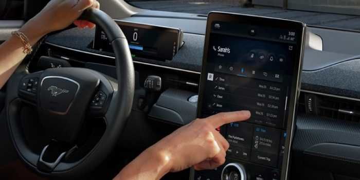 A driver holding the steering wheel of a Ford Mustang Mach-E uses her index finger to navigate the large vertical infotainment touchscreen.