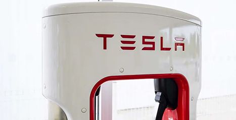A close-up of a white Tesla Supercharger station with the red Tesla logo at the top and a red-trimmed opening for the charging handle.