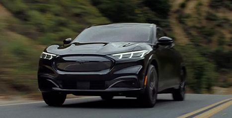 A front view of a black Mustang Mach-E driving fast on a winding road through a hilly landscape.