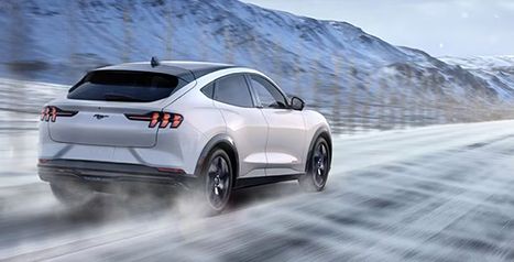 Rear view of a white Mustang Mach-E driving on a snow-covered road through a wintry mountain landscape.