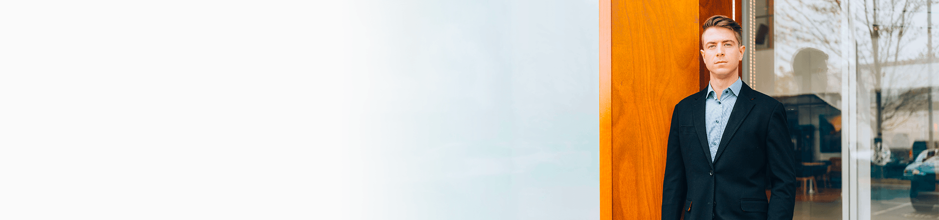 A wide banner image featuring a man in a black blazer on the right, leaning against a wooden pillar outside a dealership. The left side fades into a solid white background.
