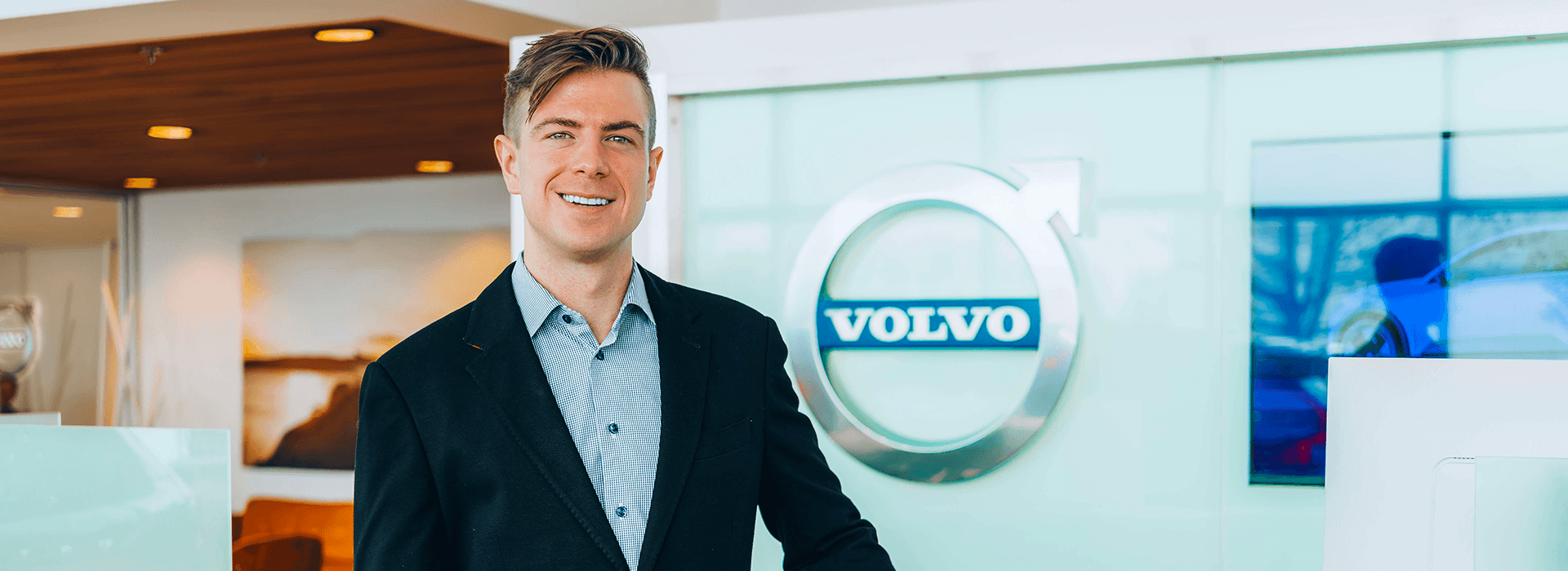 A smiling man in a black blazer and patterned dress shirt standing in front of a Volvo logo at a dealership.