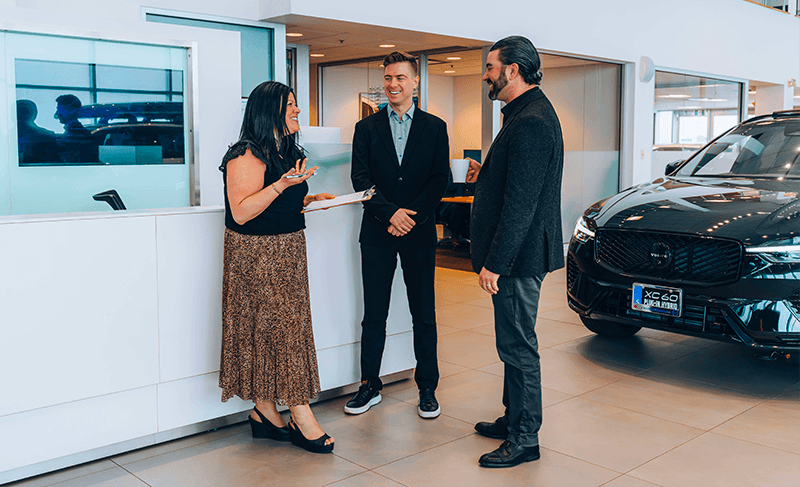 Three dealership staff members smiling and talking on the showroom floor next to a black Volvo XC60.